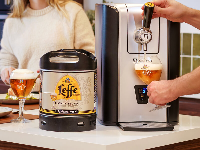 A PerfectDraft machine, pouring a Leffe Blonde beer into a Leffe glass, with a keg of Leffe Blonde sat beside it.