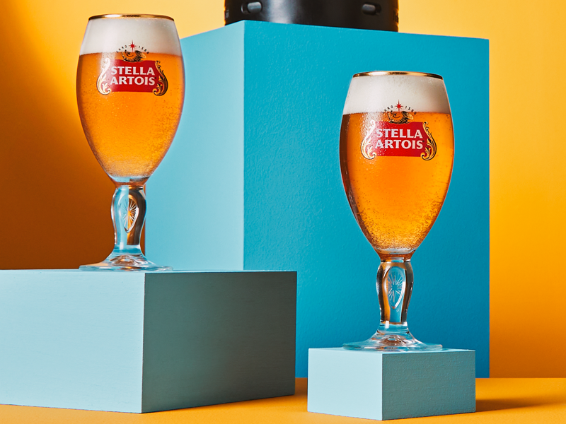 A picture of two Stella Artois chalices filled with Stella Artois beer, stood on two light blue blocks with an orange background.